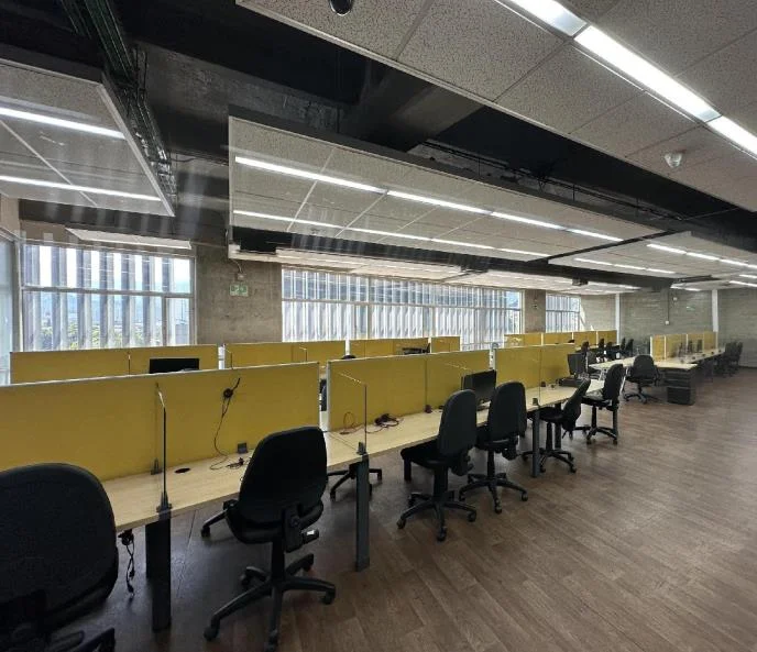 Puestos de trabajo con divisores amarillos y sillas negras, piso de madera y ventanales de piso a techo.