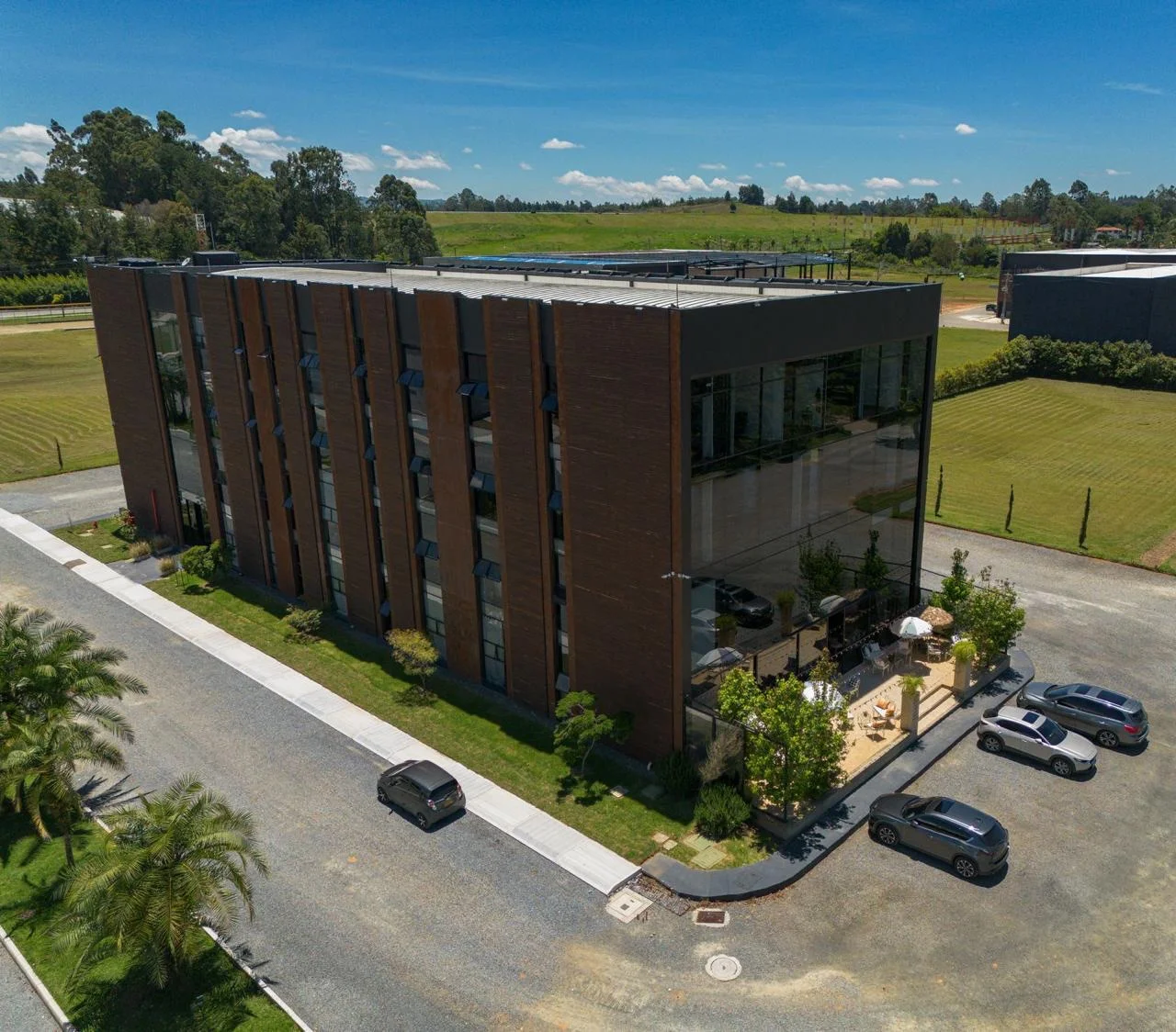 Vista aérea del edificio corporativo de fachada en acero corten y vidrio, con parqueadero, zonas verdes y una terraza con mobiliario exterior en la esquina.