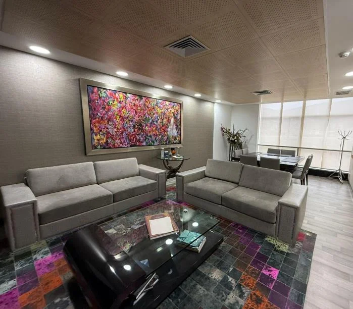 Sala de estar con sofás grises, una pintura abstracta de gran formato y un comedor al fondo junto a la ventana.