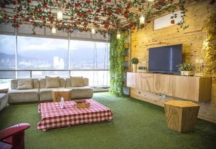 Sala de descanso con instalación floral en el techo, sofás claros, pared de madera con televisor y vista panorámica de la ciudad.