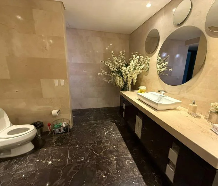 Baño con paredes de mármol claro, piso oscuro, espejos redondos superpuestos y un arreglo floral blanco.