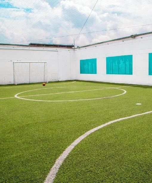 Cancha de fútbol techada con césped sintético, arco y balón, y paneles decorativos color turquesa en las paredes.